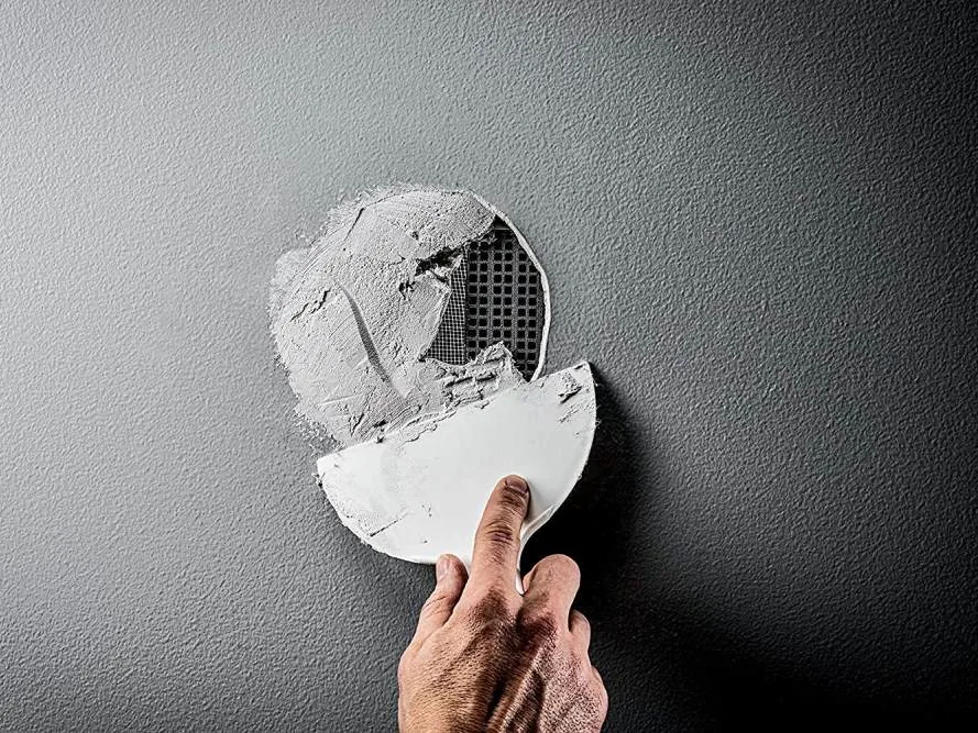 A hand using a putty knife to apply joint compound over a mesh patch in a wall