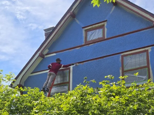 A painter on a ladder painting the blue exterior siding and trim of a two-story house