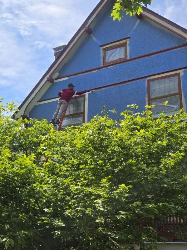 A painter on a ladder painting the blue exterior siding and trim of a two-story house