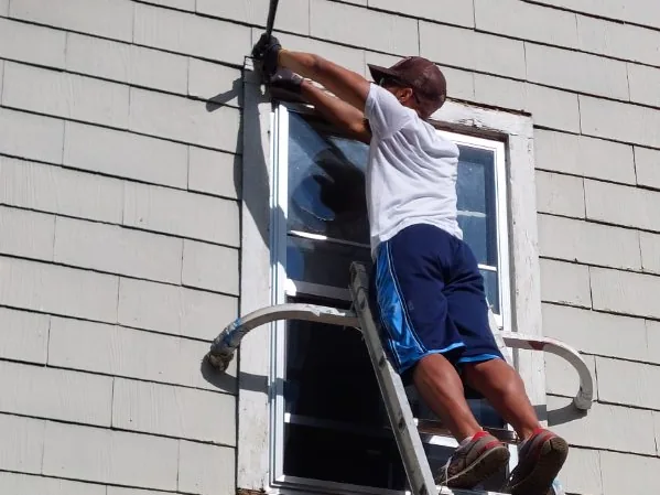 A painter on a ladder working on a window frame on a house with gray siding