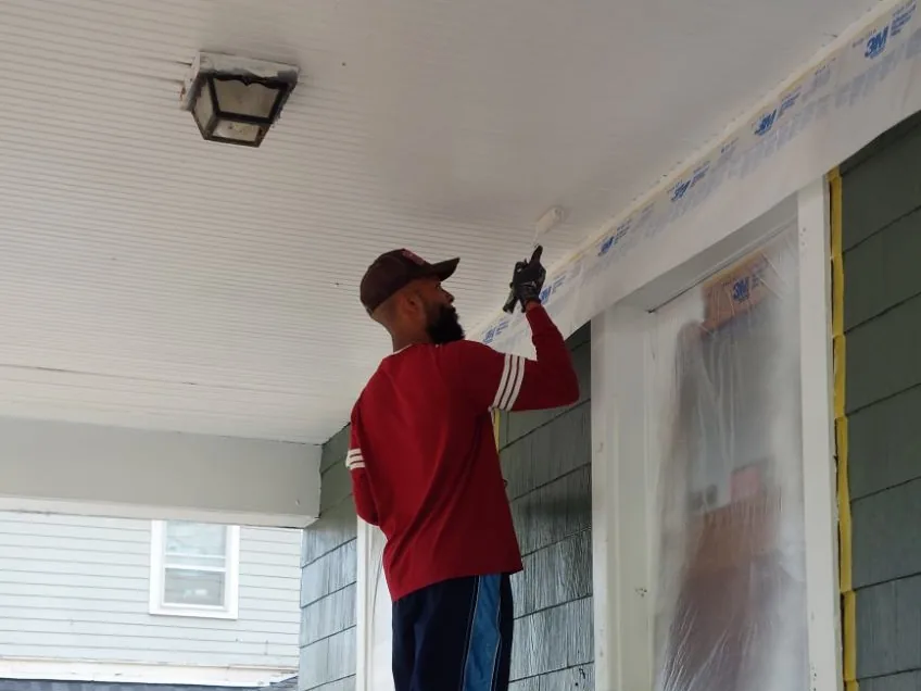 A painter working on the ceiling and trim of a taped and masked front porch