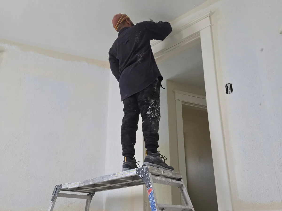 A painter on a platform ladder painting the ceiling edge and trim in a primed room